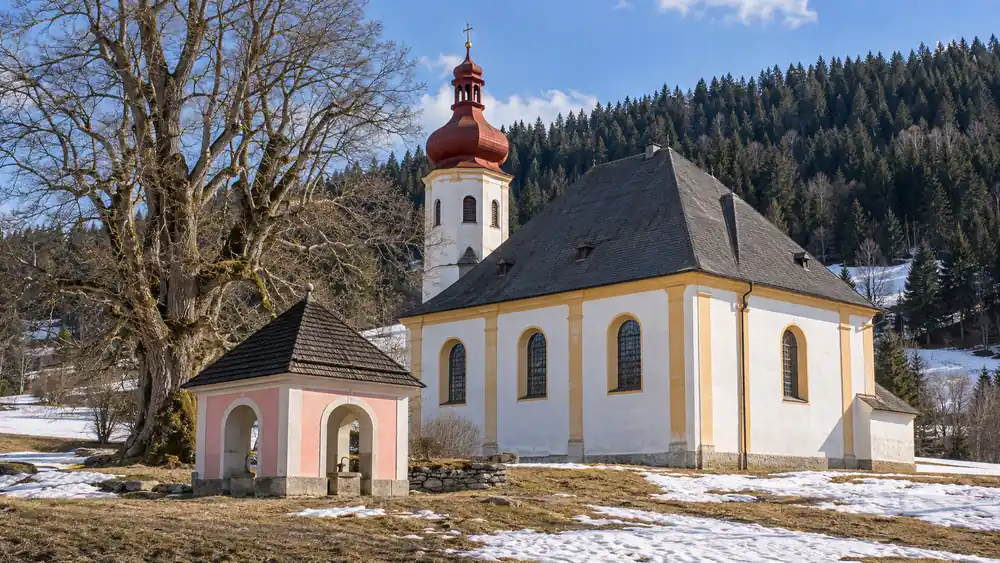 Bílý barokní kostel s věží zakončenou červenou cibulovou bání, před ním růžová kaplička nad pramenem a stará lípa