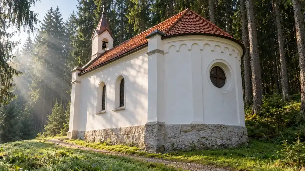 Malá bílá kaple s červenou taškovou střechou, zvoničkou a kulatým oknem v oblé apsidě pod vysokými smrky v ranních paprscích