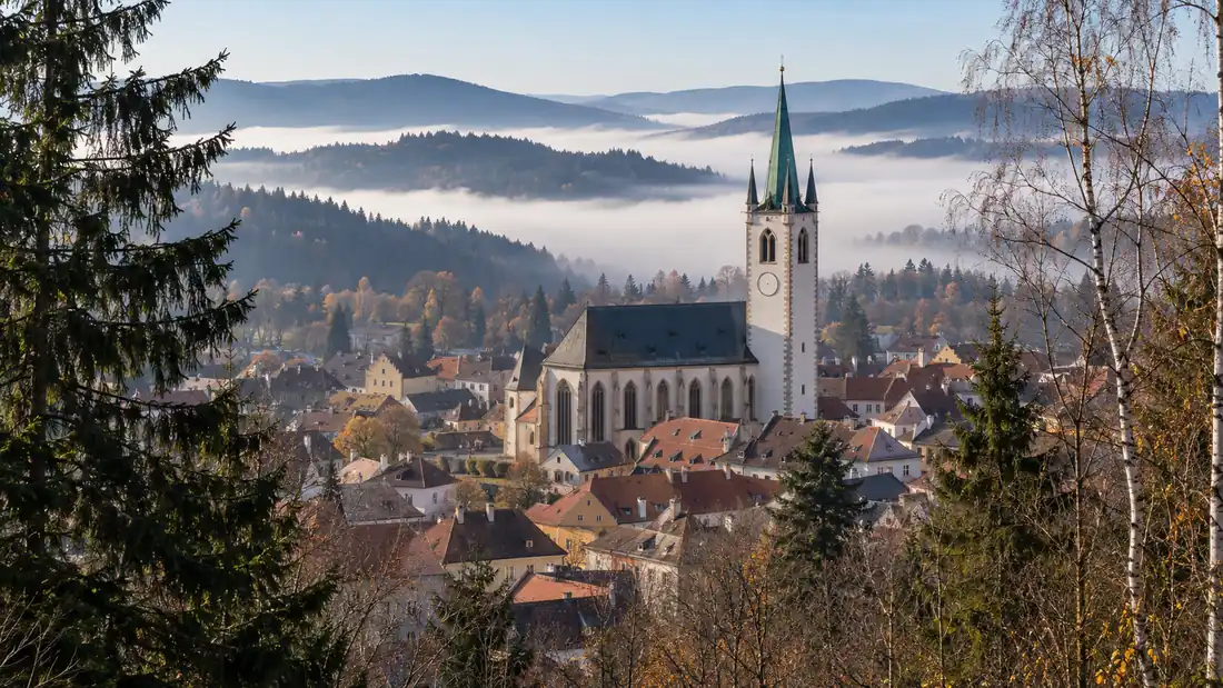 Pohled přes střechy horního města na vysokou bílou kostelní věž se zelenou střechou, za městem ranní mlha v údolích