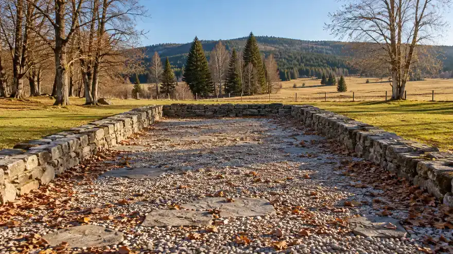 Nízké kamenné zídky obrysu zbořeného kostela vysypané oblázkovým štěrkem se spadaným listím, kolem louka, holé stromy a smrky v podzimním slunci