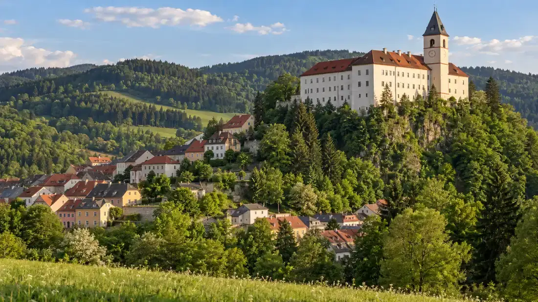 Město Vimperk na zelených svazích nad údolím a nad ním na zalesněném skalním ostrohu dlouhá bílá budova zámku se štíhlou hodinovou věží